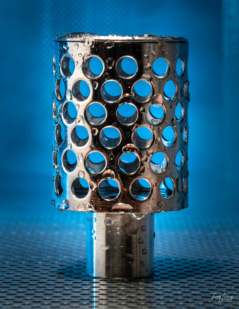 A sink trap with water on it, photographed with a blue background on steel.