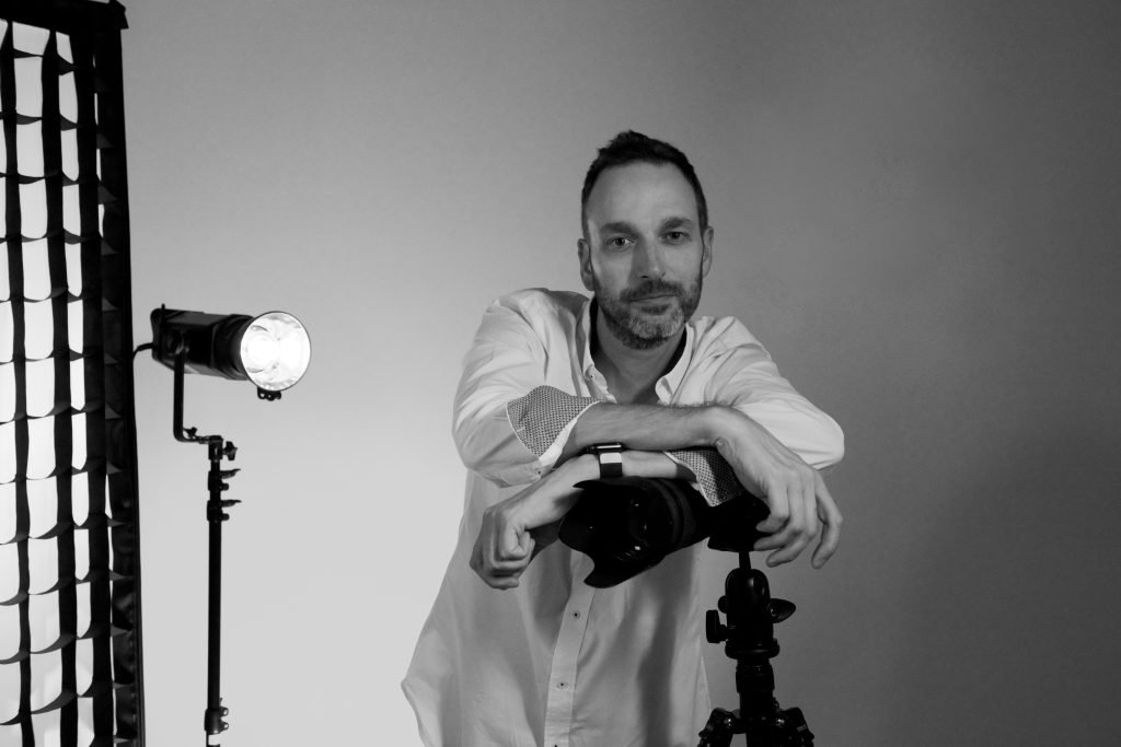 Terry Livesey in the studio resting on the camera which is on a tripod. To the left is a light and strip softbox.