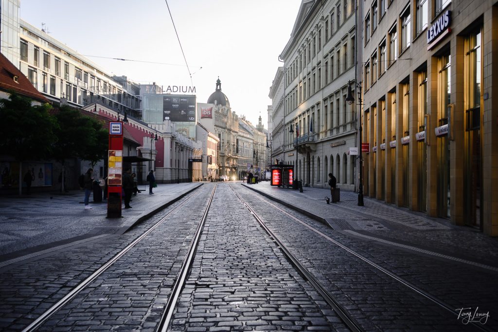 A quiet cobbled street with tram tracks winds between historic Prague buildings at sunrise; a few people stand on the pavements and shopfronts line the street.