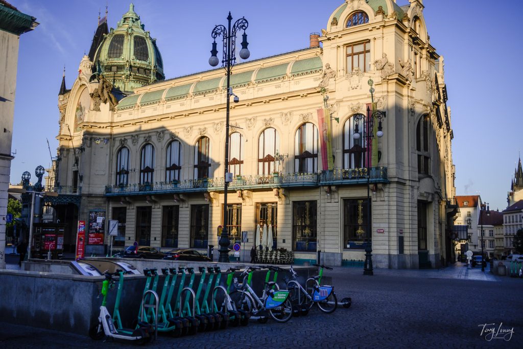 A historic building with ornate architecture and arched windows stands in sunlight in Prague. In the foreground, rows of green hire bikes and scooters are parked on a cobblestone plaza.