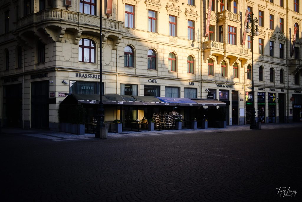 A sunlit European street in Prague with a large, ornate yellow building housing a brasserie, café, bar, and wine & spirits shop; outdoor seating is arranged under black awnings on a cobbled plaza.