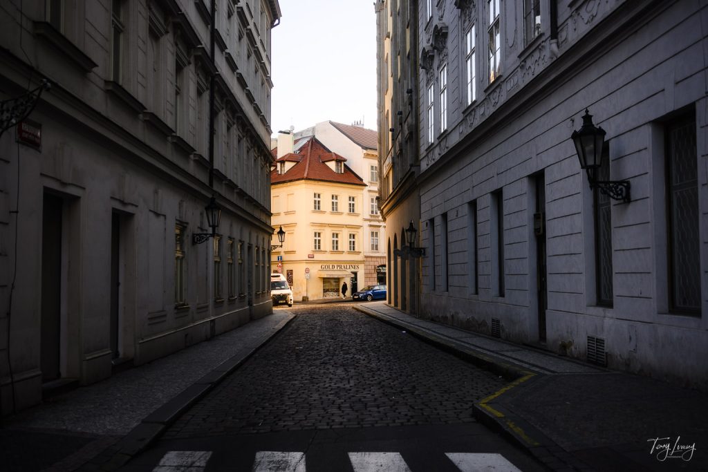 A narrow cobblestone street in Prague, lined with tall, old buildings, leads to a sunlit square with cream-coloured architecture and a sign reading Old Prague. Traditional street lamps hang on the building walls.