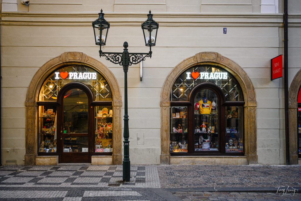 Two arched shop windows display souvenirs under neon I ❤️ PRAGUE signs on a cobbled street in Prague, with a vintage streetlamp standing before the cream-coloured building facade.
