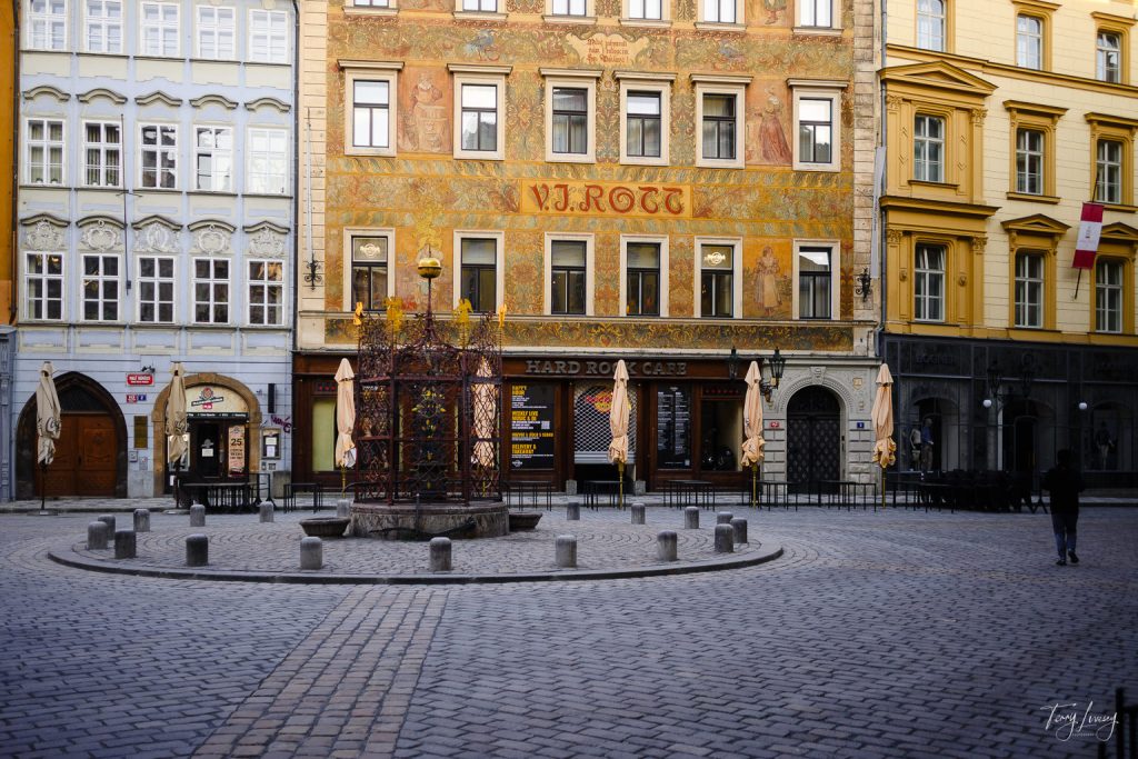 A cobblestone square in Prague with a decorative iron fountain in the centre, surrounded by colourful historic buildings, including one with a “V.J. ROTT” sign and a Hard Rock Café on the ground floor.