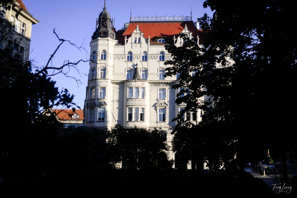 A grand historic building in Prague with a red-tiled roof, ornate white façade, and towers, partially framed by trees casting shadows in the foreground on a sunny day.