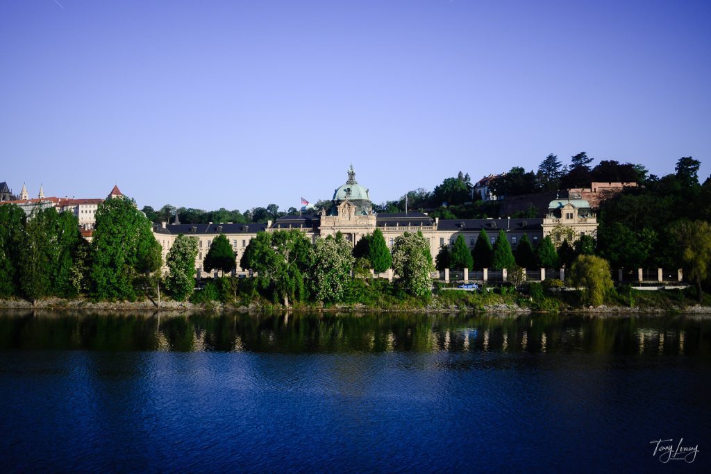 A grand historic building with a dome and surrounding greenery sits across a calm river in Prague, under a clear blue sky.
