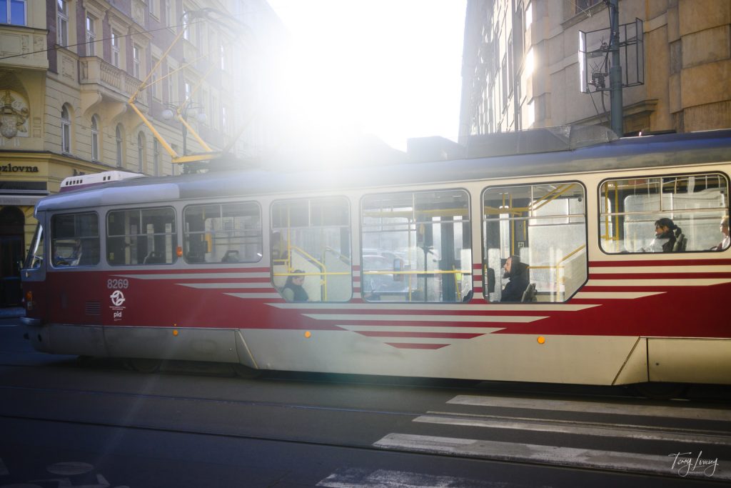 A red and white tram with passengers inside travels through a Prague city street, illuminated by bright sunlight. The buildings and shops in the background suggest an urban setting.