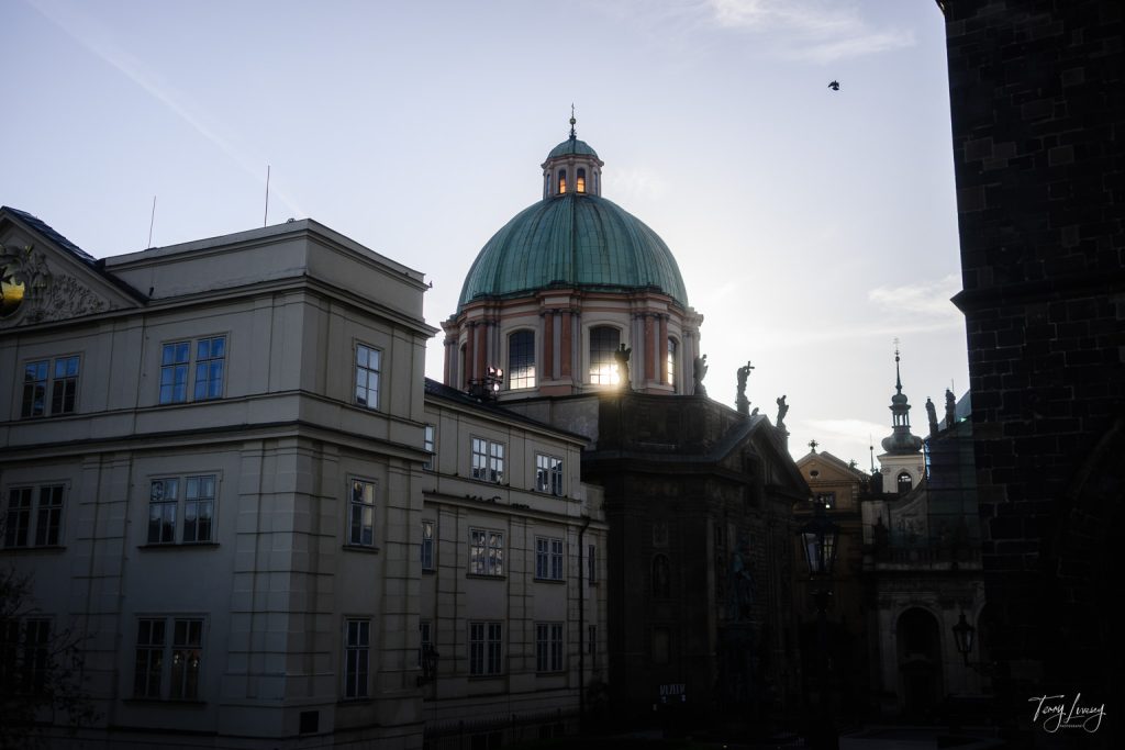 A domed building with statues on its roof stands against a bright Prague sky at sunset, partially obscured by nearby classical-style buildings; a single bird is flying in the sky.