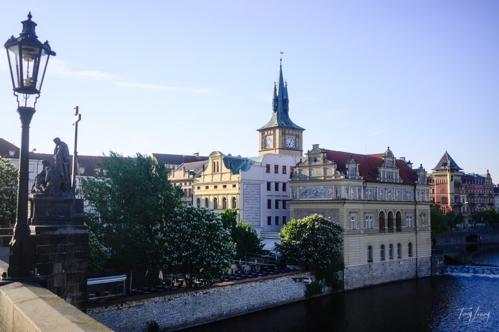 Historic buildings with ornate façades and a tall clock tower line the river in Prague, all beneath a clear blue sky, seen from a bridge adorned with a lamp-post and statue on the left.