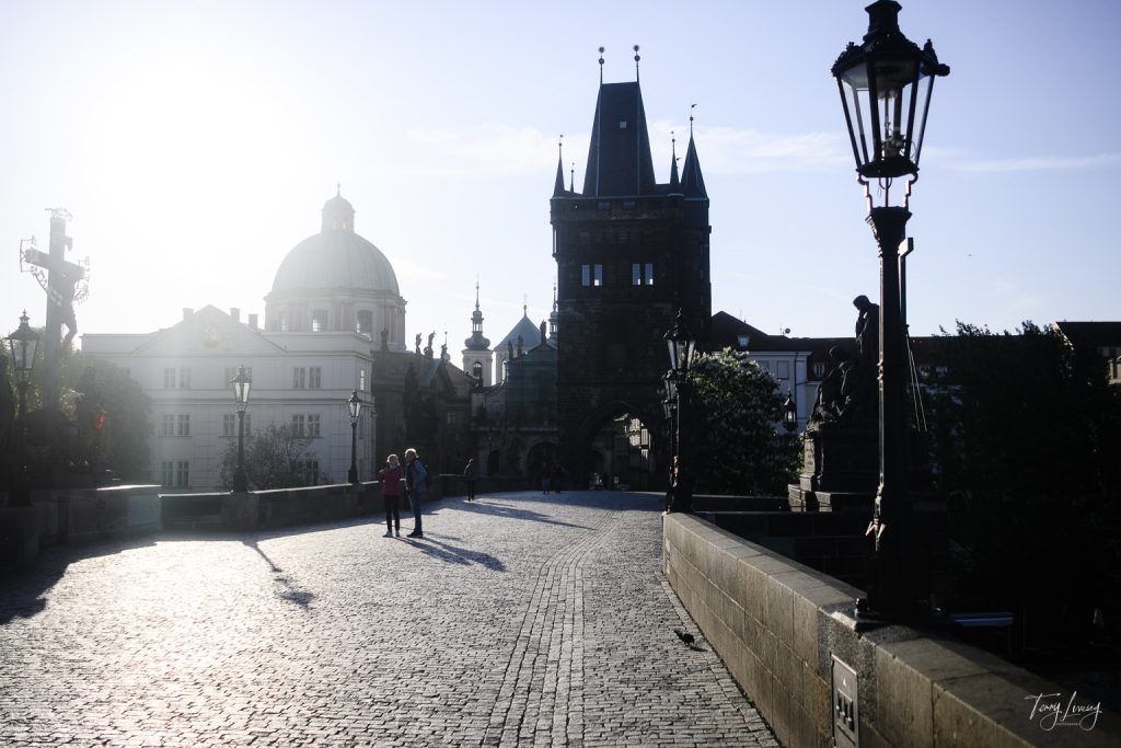 A cobblestone bridge in early morning light with a few people walking, historic Prague buildings, and ornate street lamps; a large tower stands at the end of the bridge silhouetted against the bright sky.
