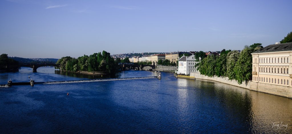 A wide river flows through Prague, with historic buildings lining the right bank, leafy trees, and several stone bridges crossing the water under a clear blue sky.