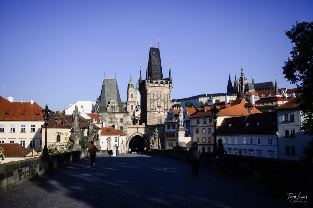 A view of Charles Bridge in Prague, with people walking, historic stone towers and statues, and red-roofed buildings beneath a clear blue sky captures the charm of this iconic Prague landmark.