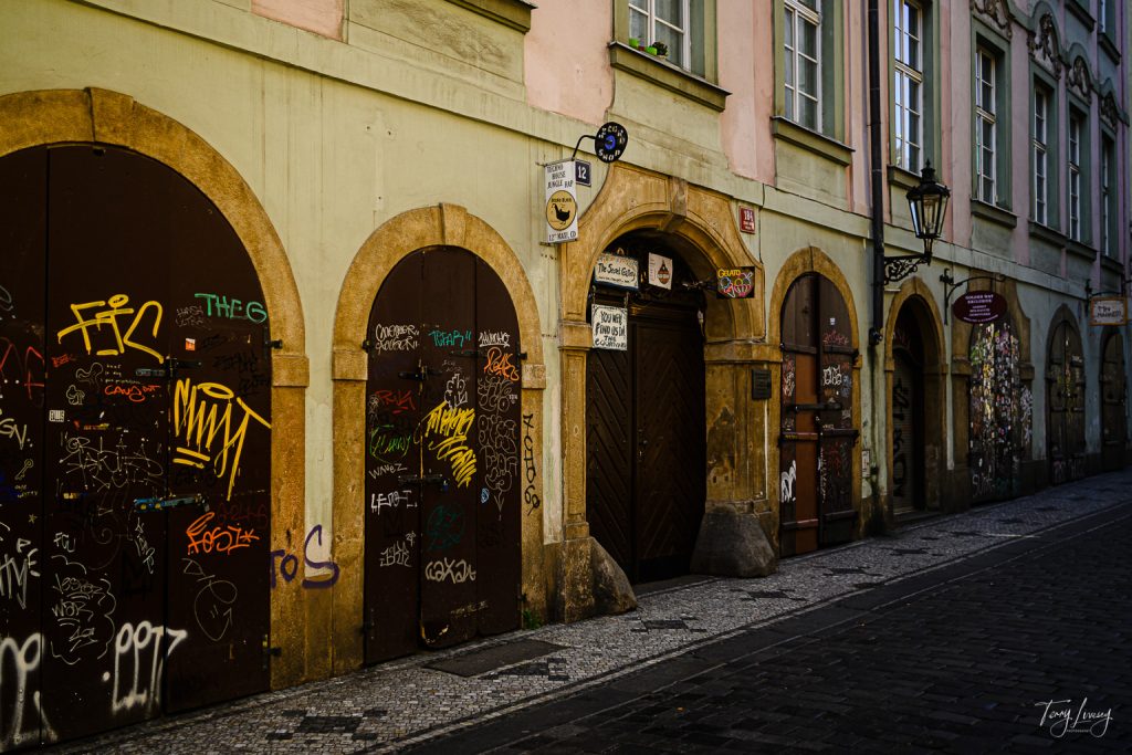 A narrow cobblestone street in Prague with old buildings featuring arched doorways covered in colourful graffiti, wooden doors, and upper-storey windows with flower boxes. The street is empty and lit by natural daylight.