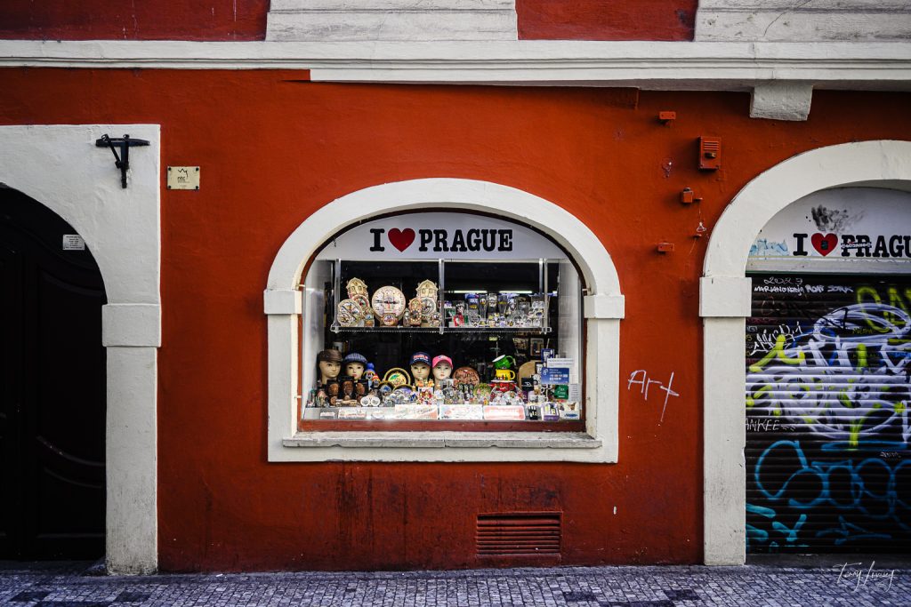 A souvenir shop with a bright red exterior displays Prague trinkets and I ♥ PRAGUE signs in the window, flanked by graffiti on a glass door and arched white trim around the window and entrance.