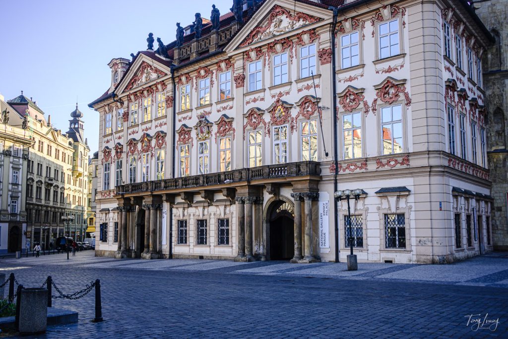 A grand, ornate building with a detailed white and red façade, large windows, and arched entrance stands on a quiet cobbled square in historic Prague.