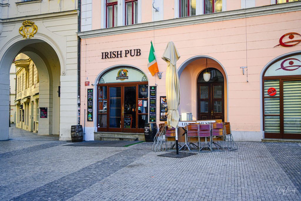 An Irish pub with a sign, Irish flag, chalkboards, and outdoor seating with stacked chairs and a folded umbrella lines a cobbled street in historic Prague.