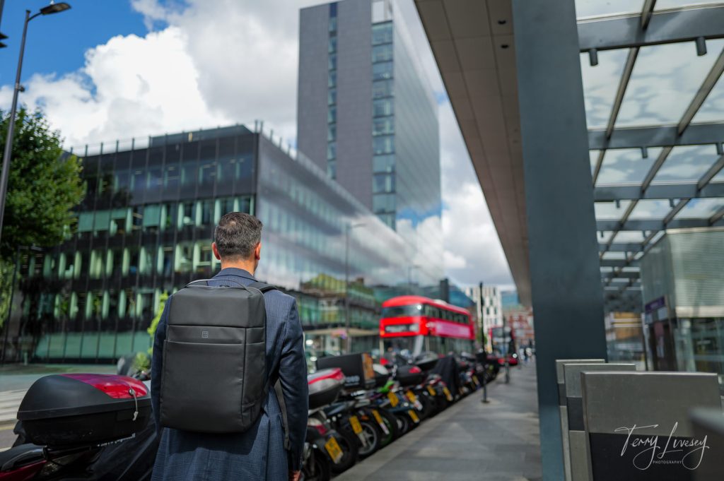 A male model walking down the street with a PW3R backup on with a red London bus comping towards him