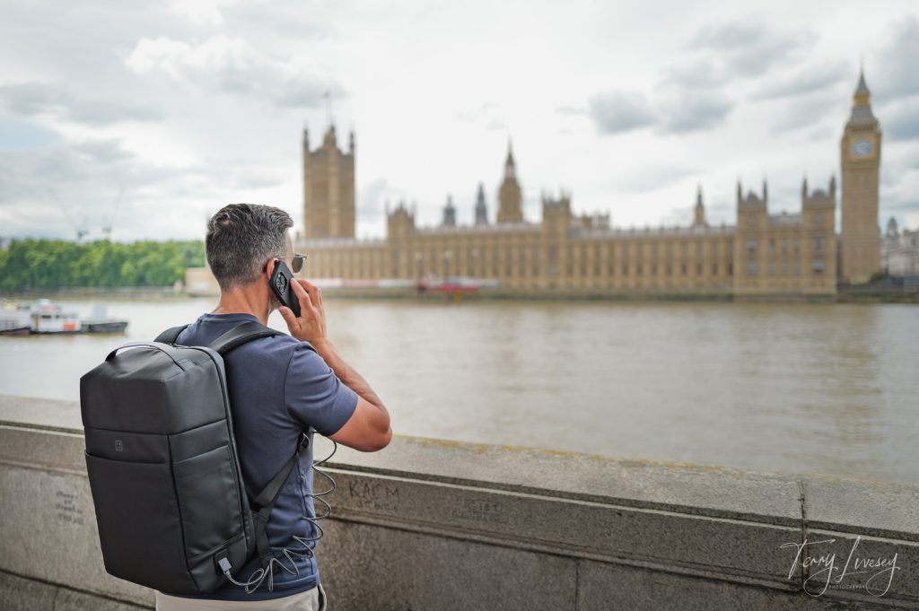 Male model holding a mobile phone connected to a backpack in front of the Houses of Parliament, London