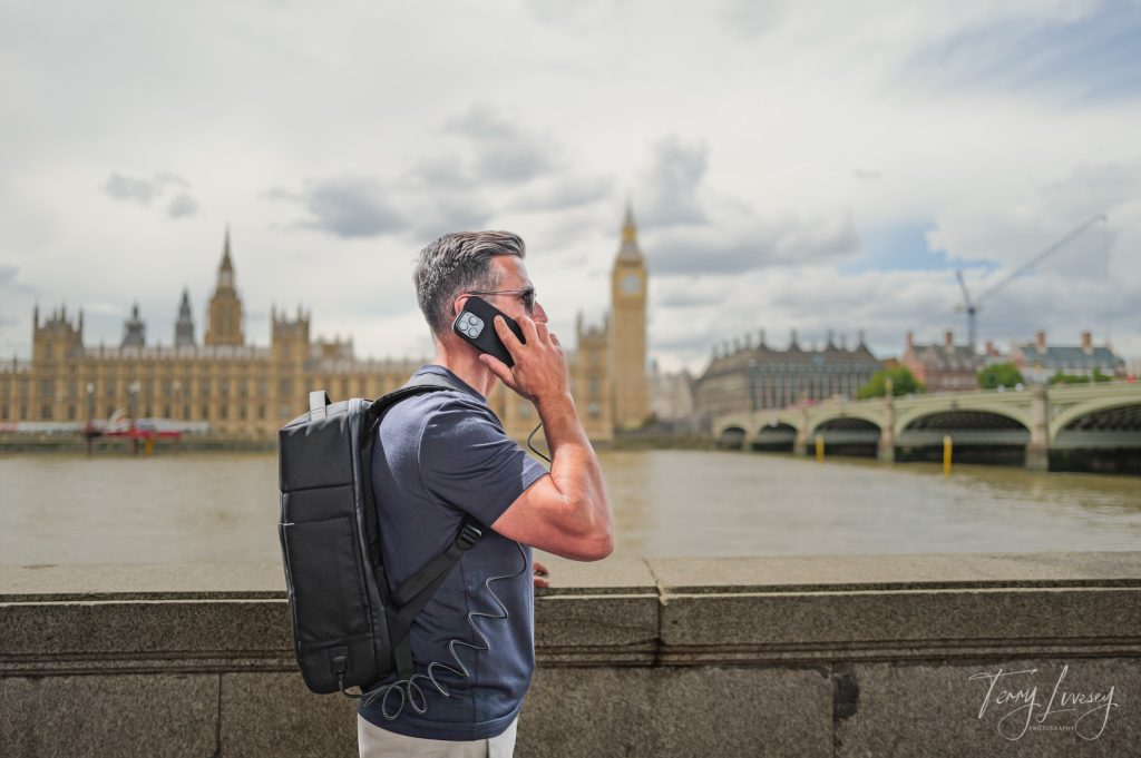 Male model holding a mobile phone connected to a backpack in front of the Houses of Parliament, London