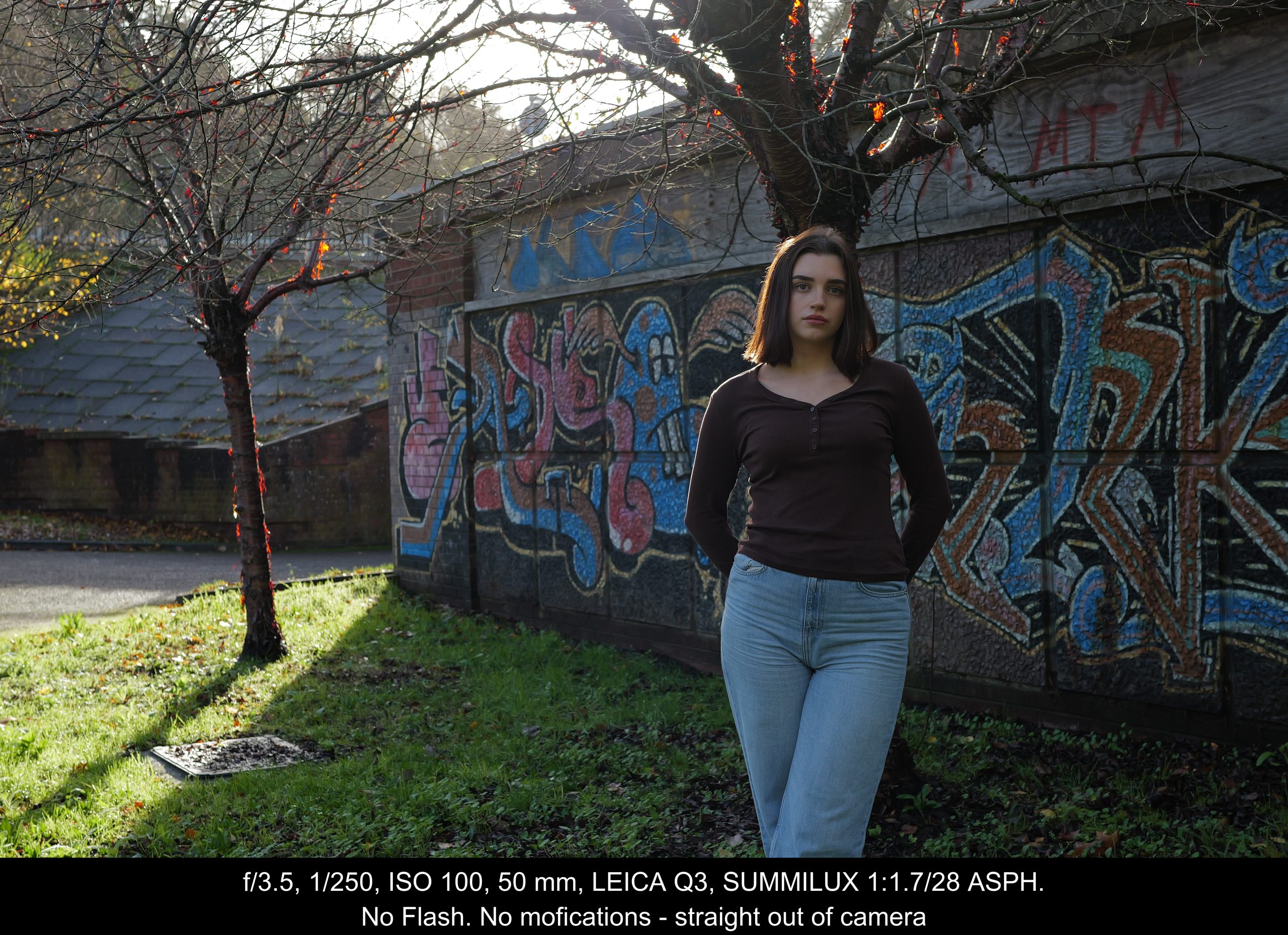 A woman with straight dark hair, wearing a brown top and blue jeans, stands in front of a graffiti-covered wall outdoors in sunlight. Leafless trees and a building are in the background.