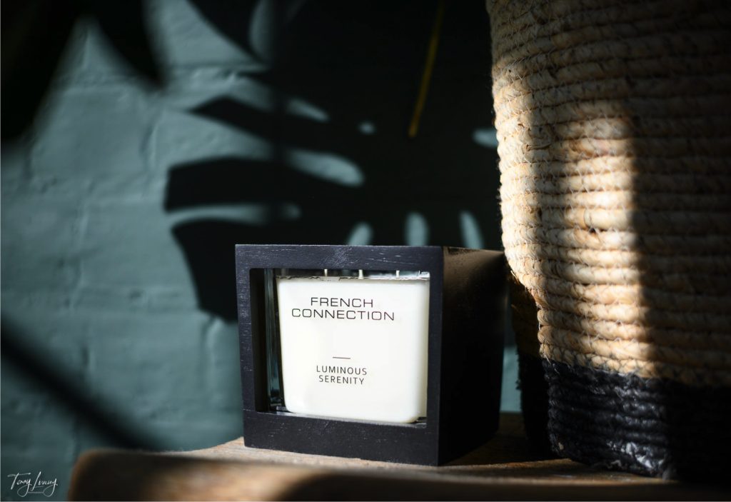 A white French Connection candle labelled Luminous Serenity rests in a black holder beside a woven basket, with sunlight casting gentle shadows on the green wall behind them.
