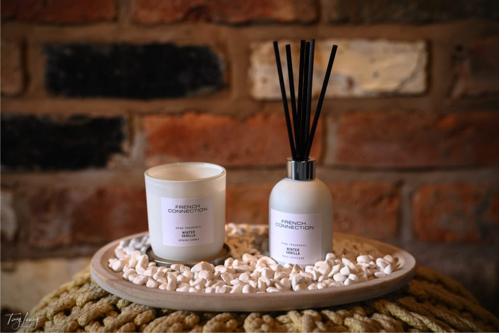 A French Connection candle and reed diffuser sit on a round tray filled with white pebbles, set against a rustic brick wall backdrop.