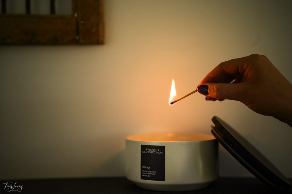 A hand with dark-painted nails lights a match to ignite a large white French Connection candle labelled Refresh, creating a cosy ambience on a dark surface against a light-coloured wall.