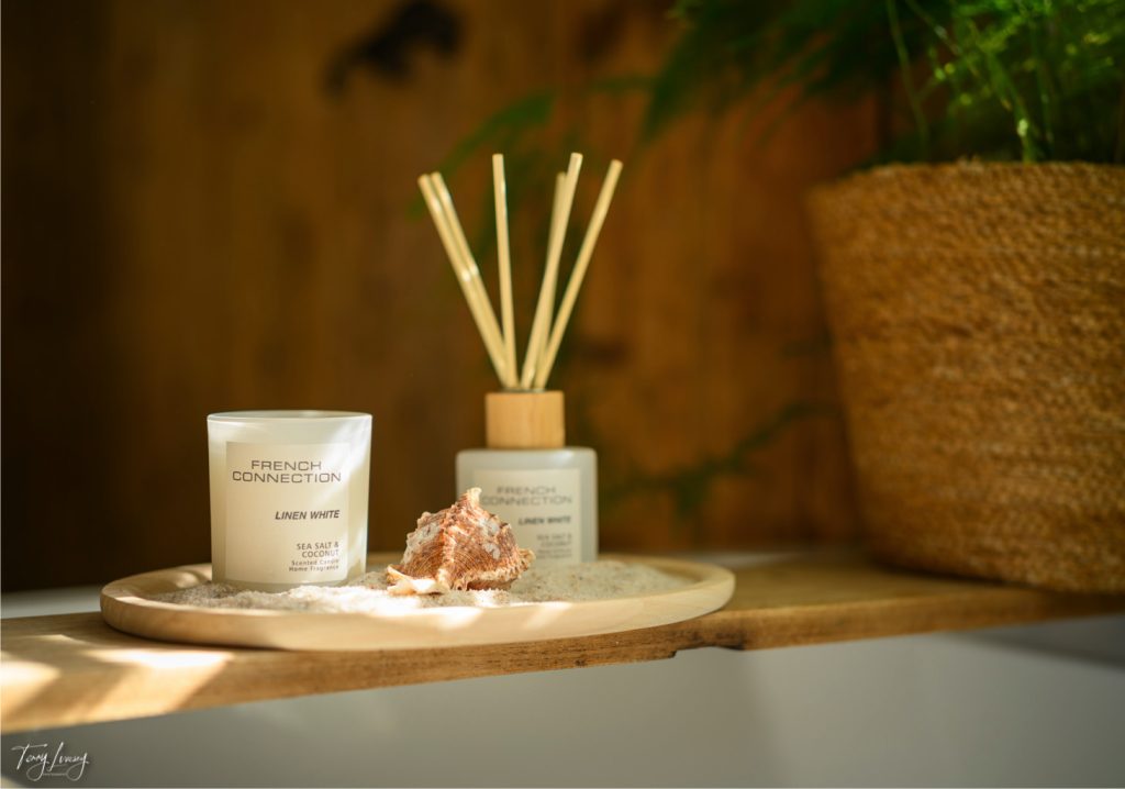 A tray with a candle, reed diffuser, and seashell from French Connection sits on sand, next to a woven basket with a green plant. Warm, natural light highlights the cosy, spa-like setting.