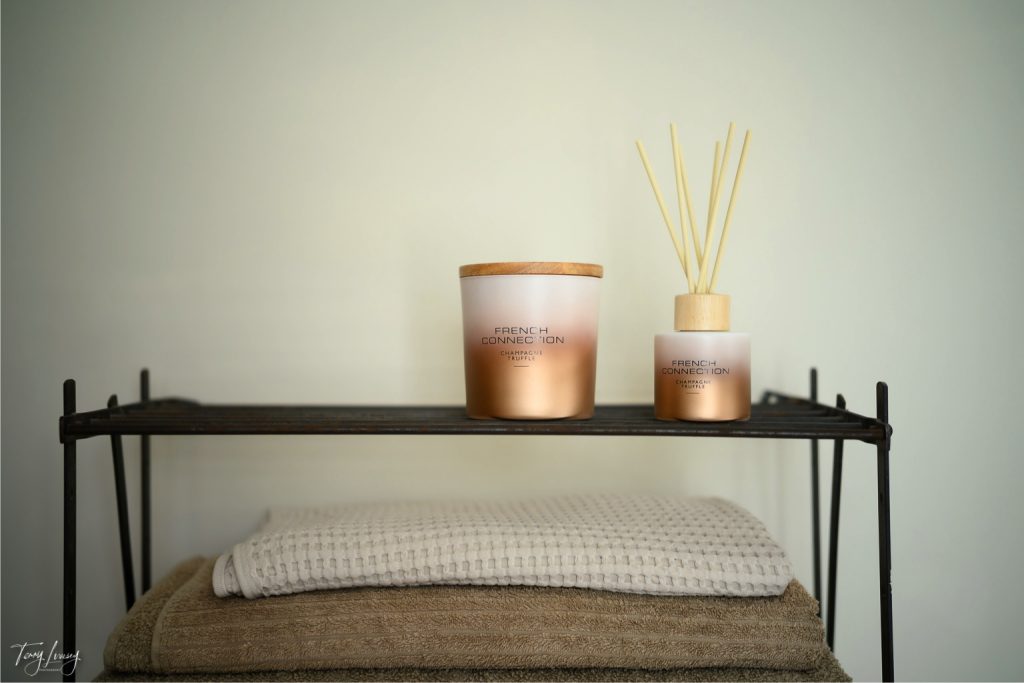 A shelf with folded beige towels, a French Connection gold candle with a wooden lid, and a matching French Connection reed diffuser in a frosted glass bottle against a plain light wall.