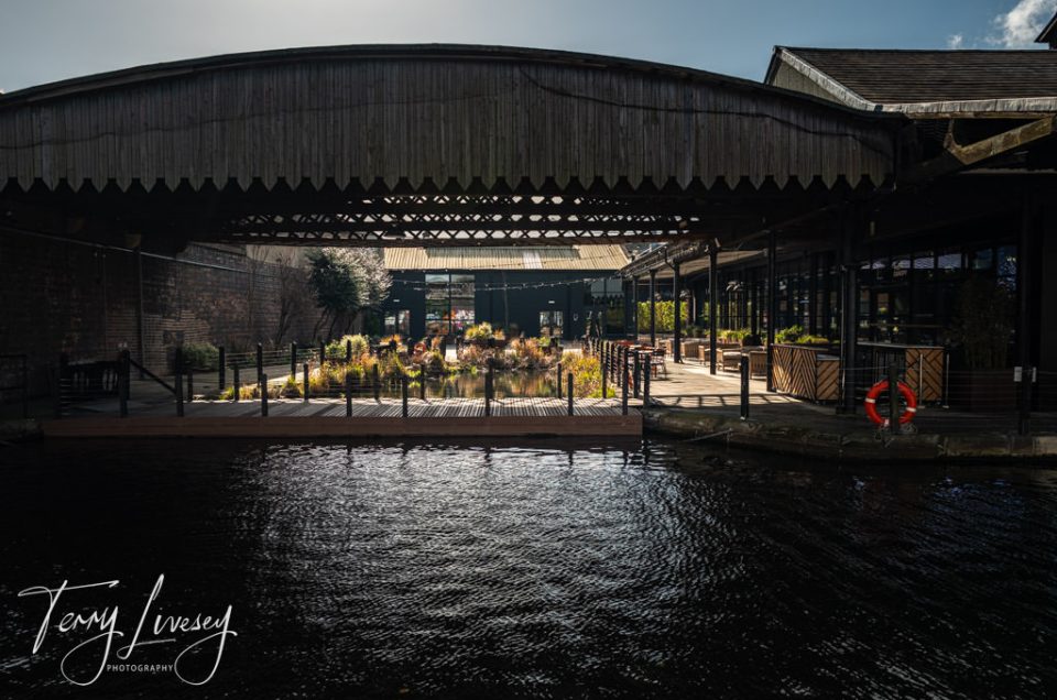 A well-lit outdoor seating area by a canal, underneath a large canopy structure, in Birmingham, United Kingdom.