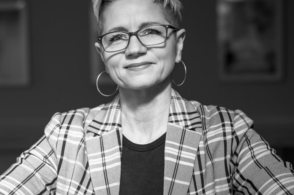 A confident woman with short hair and glasses, wearing a checked blazer and hoop earrings, sits with arms folded and smiles slightly in a black-and-white portrait. Blurred frames are visible in the background.
