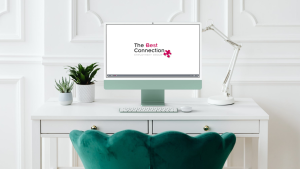 A modern white desk with a green chair, a desktop computer displaying The Best Connection Employment Group logo, a white lamp, a keyboard, mouse, and two potted plants in a bright room.