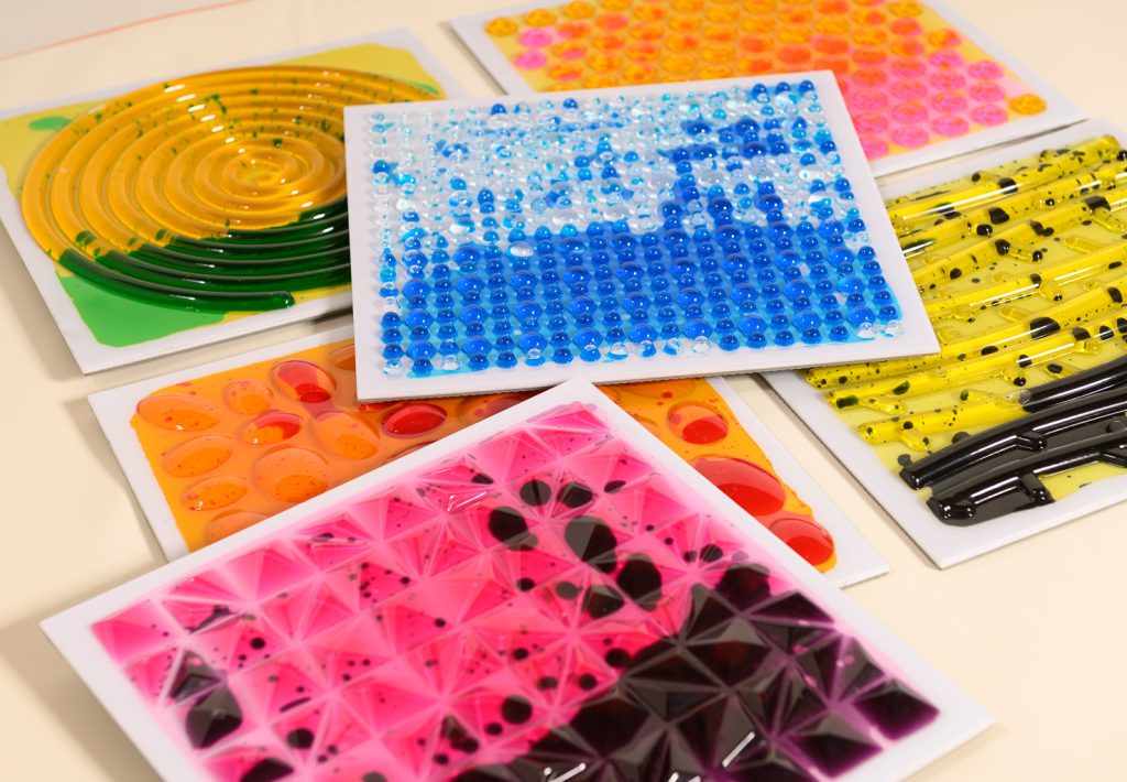 Five rectangular tactile art pieces with raised textures and vibrant colours, including swirls, dots, and geometric shapes in yellow, blue, pink, orange, black, and white, are arranged on a flat surface.