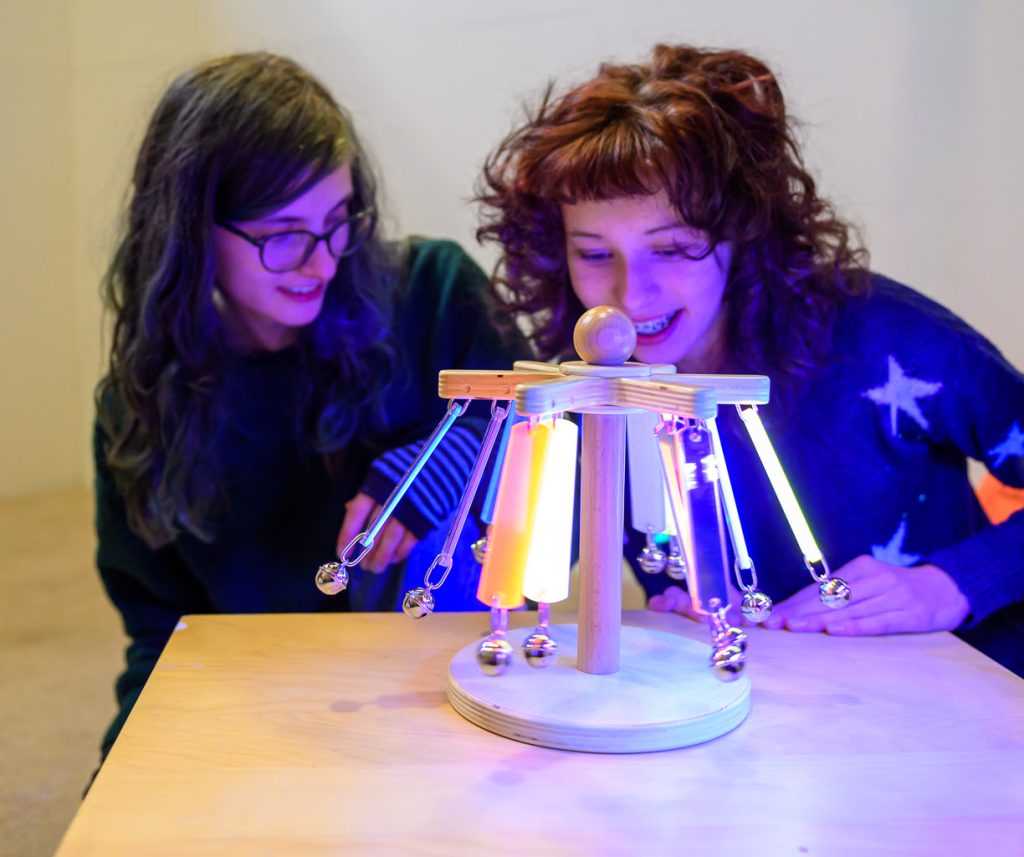 Two people with long hair sit at a table, smiling and looking at a wooden structure with hanging colourful glowing rods and small bells. The room is warmly lit, and the atmosphere appears playful and curious.