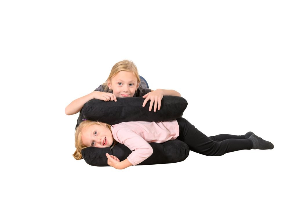 Two young girls are playing and smiling, one lying on the floor and the other leaning over her, both surrounded by large black cushions, against a white background.