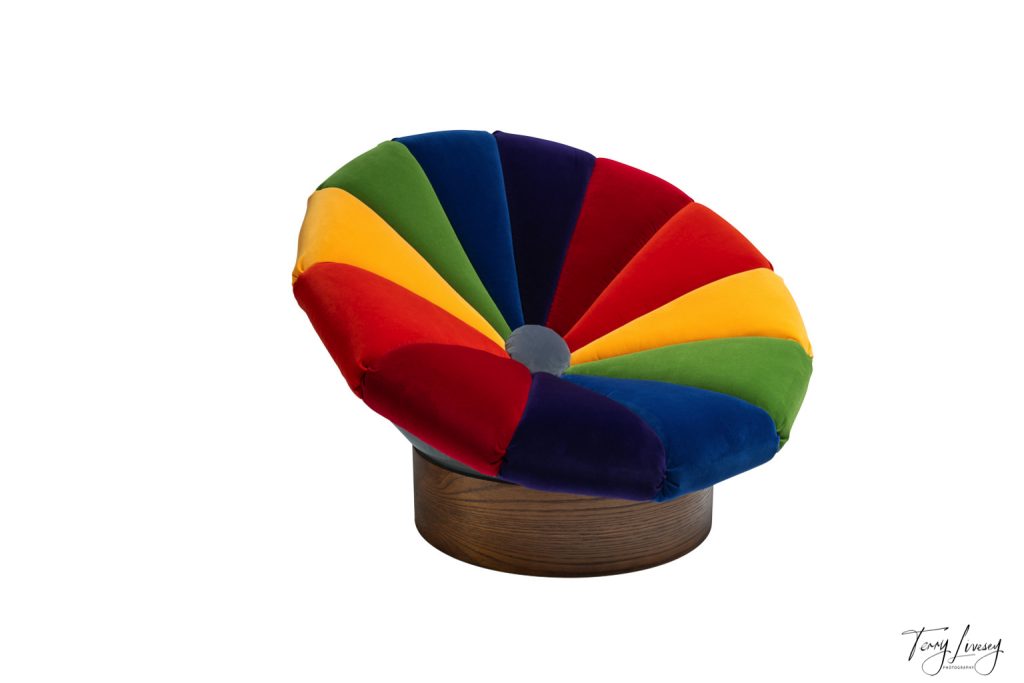 A round, cushioned chair with a wooden base, featuring a vibrant, rainbow-coloured upholstery in alternating segments of red, yellow, green, and blue, set against a plain white background.