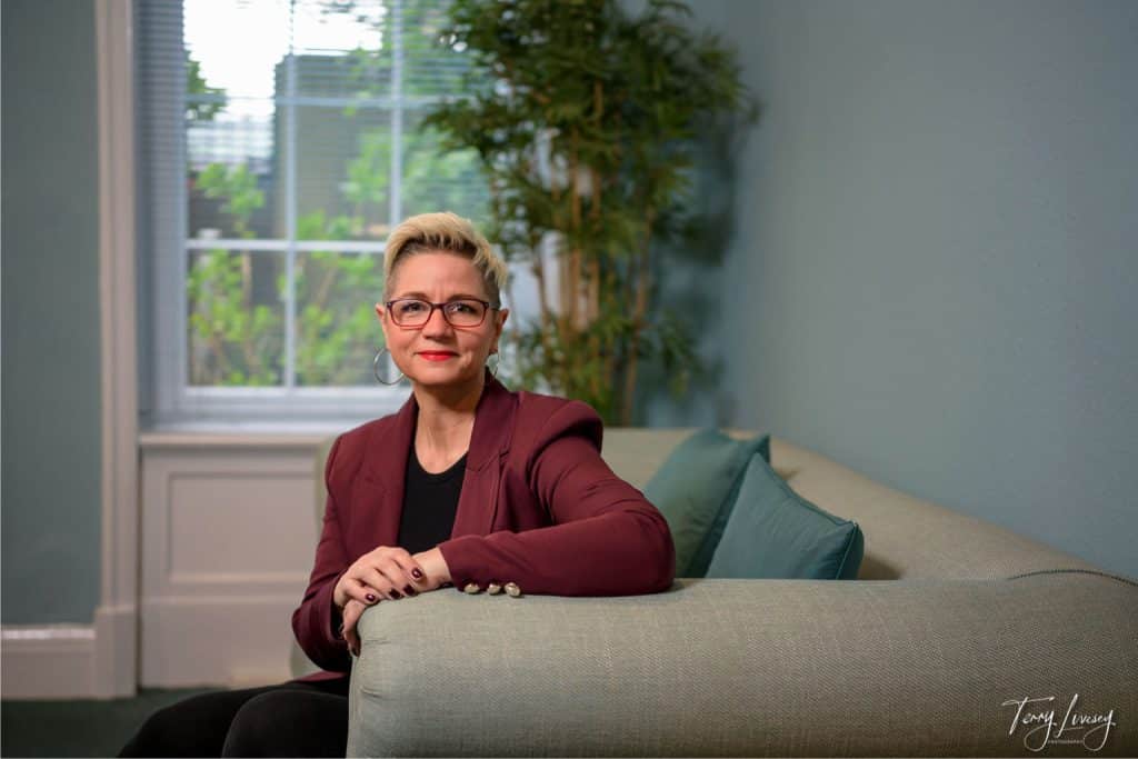 A person with short blond hair and glasses, wearing a maroon blazer, sits on a light grey sofa in a softly lit room with a large window, green plant, and blue walls—an ideal setting for personal brand photography Worcester.
