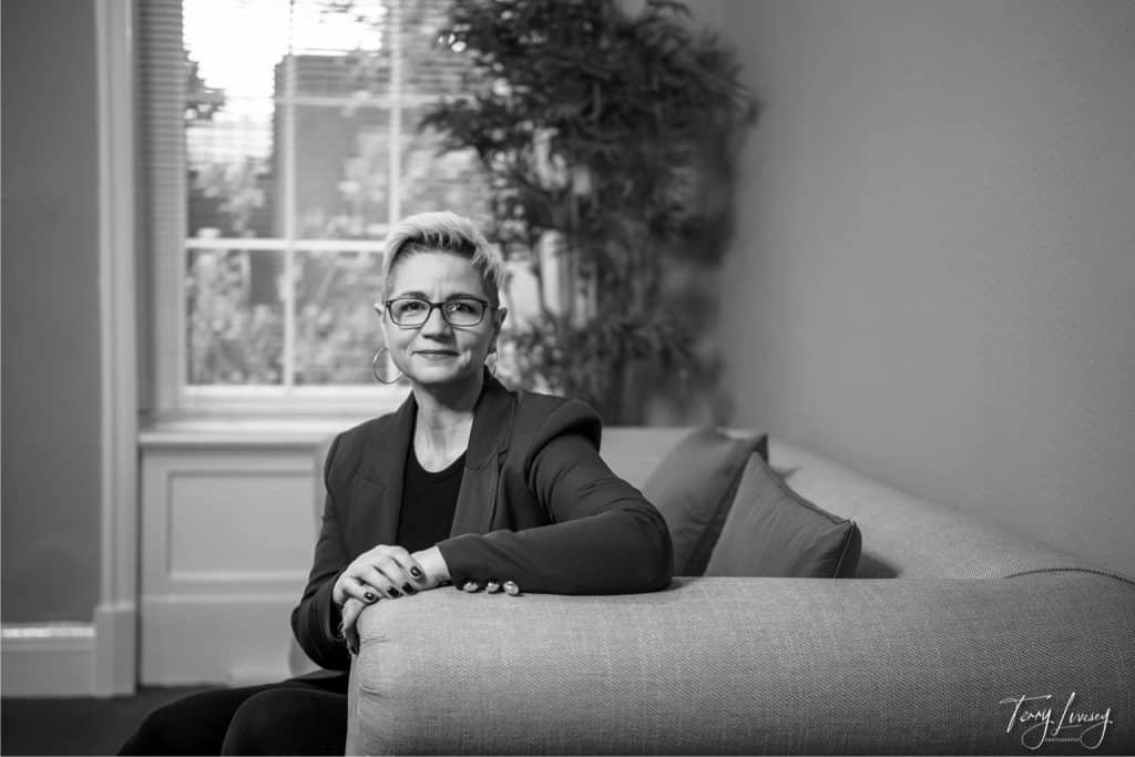 Black and white photo of a person with short, fair hair and glasses, sitting on a sofa with hands resting on the armrest, in a room with a large window and potted plant—ideal for personal brand photography in Worcester.