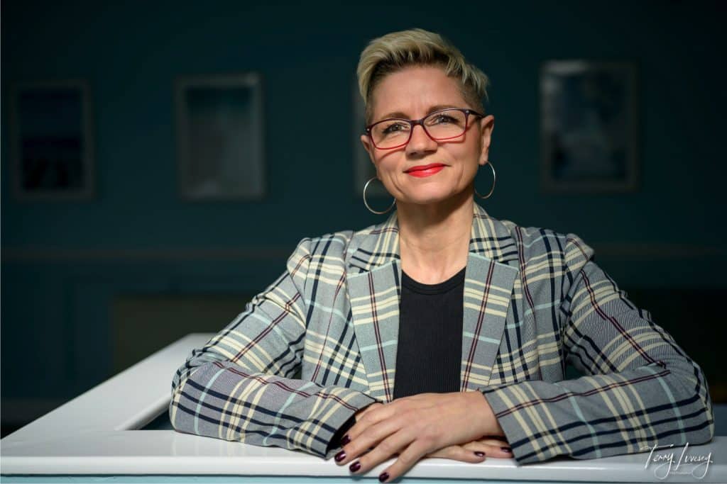 A confident woman with short blonde hair, glasses, and hoop earrings, wearing a checked blazer and black top, smiles while resting her arms on a white surface—a perfect example of personal brand photography Worcester—against a blurred blue background.