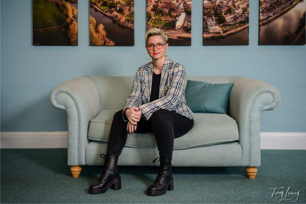 A person with short blonde hair, glasses, and red lipstick sits on a light blue sofa—an ideal setting for personal brand photography in Worcester—wearing a checked blazer, black trousers, and boots. Framed aerial city photos hang on the blue wall behind them.